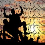 Silhouetted people climbing barbed wire fence sunset background