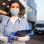 Nurse in scrubs and mask outside hospital holding clipboard