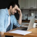 Man stressing over work on laptop at kitchen table.