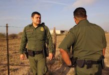 Border Horror: Convicted Felon Caught Sneaking In! Two uniformed officers standing outdoors near a fence.