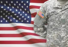 Soldier in camouflage uniform standing before American flag.