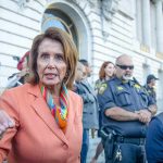 A woman in an orange blazer speaking passionately at a rally with security personnel in the background