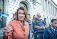 ICE Targets Pelosi – Demand She STEP ASIDE! A woman in an orange blazer speaking passionately at a rally with security personnel in the background