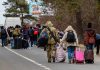 Group of people carrying luggage walking along a road with a soldier
