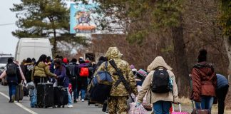 Group of people carrying luggage walking along a road with a soldier