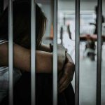 Person sitting behind bars with arms wrapped around knees.