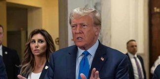 Man speaking with hands raised, woman and security behind.