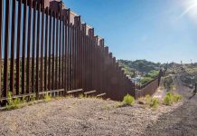Trump’s Border War ESCALATED – Militarized Zoning Begins Tall metal border wall with rural landscape.