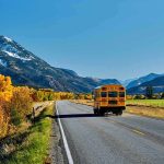 A yellow school bus driving on a rural road surrounded by autumn trees and mountains