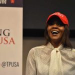 A woman wearing a red cap and a white blouse speaking at a political event