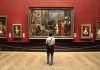 A person standing in an art gallery, observing a large historical painting surrounded by other artworks
