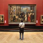 A person standing in an art gallery, observing a large historical painting surrounded by other artworks