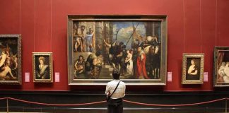 A person standing in an art gallery, observing a large historical painting surrounded by other artworks