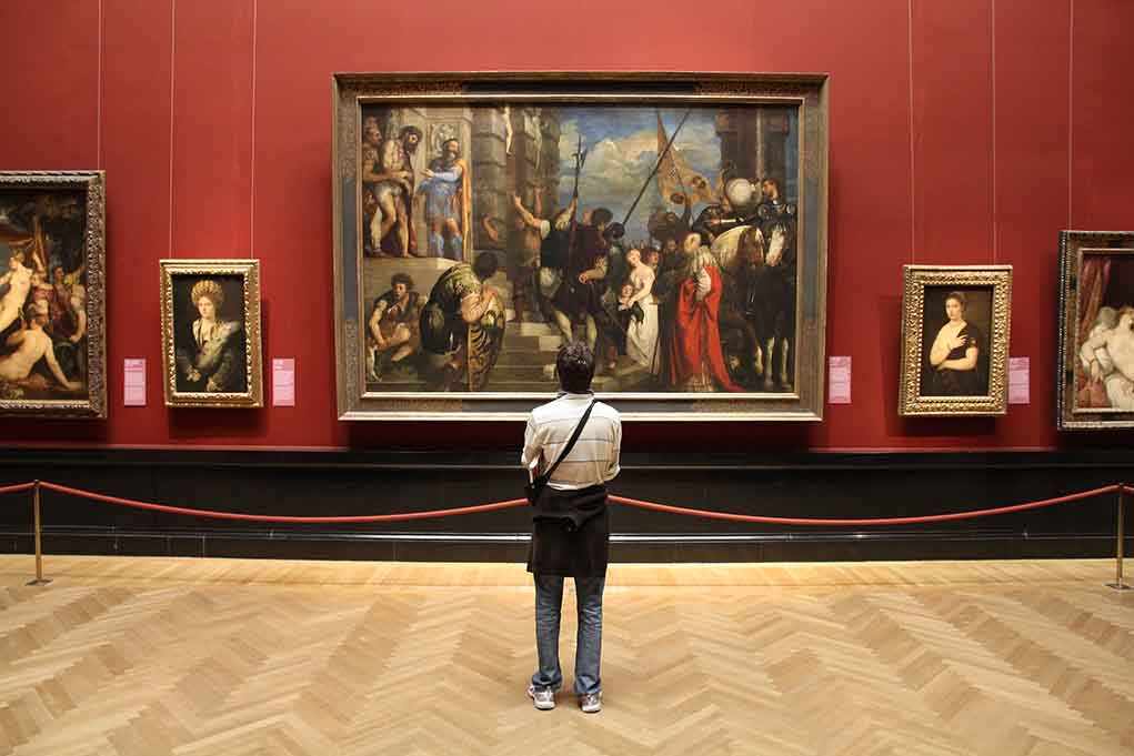 A person standing in an art gallery, observing a large historical painting surrounded by other artworks