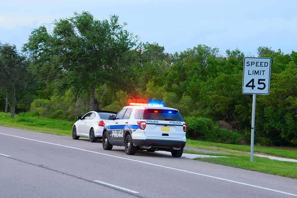 Police car with flashing lights pulling over a white vehicle beside a speed limit sign