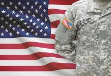 Soldier in camouflage uniform standing before American flag.