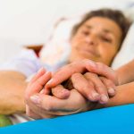 A caregiver holding the hands of an elderly patient in a hospital bed