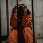 Mentally Ill Rotted in Jail—Government Shrugged Person in orange jumpsuit sitting behind bars, handcuffed.