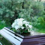 Witness to ICE Shooting Dies Mysteriously Coffin with white flowers outdoors in green surroundings.