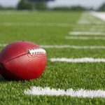 A red American football resting on a green artificial turf field