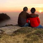 A couple sitting on a cliff watching the sunset over the ocean