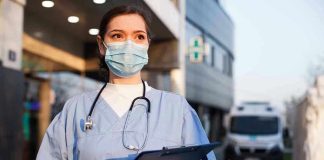 Nurse’s Disgusting Trump Threat Under Investigation! Nurse in scrubs and mask outside hospital holding clipboard.