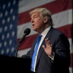 man in suit speaking at a podium with a US flag backdrop