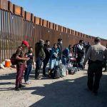 Group of people standing by a border fence with officers and vehicles