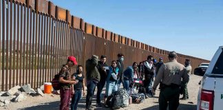 Group of people standing by a border fence with officers and vehicles