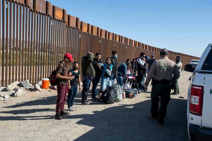 1973414822 Group of people standing by a border fence with officers and vehicles