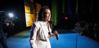 AOC Gets Brutal News As She Test-Runs Senate Bid Woman in a light suit holding a microphone on stage