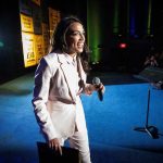 Fox News Host Taunts AOC On Live TV Woman in a light suit holding a microphone on a stage