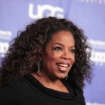 Oprah Tells Fans To CELEBRATE Murderous Mother smiling woman with curly hair on a blue step-and-repeat backdrop