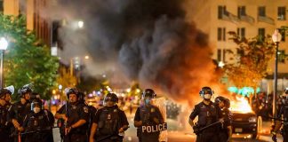 Christian Jones and Teddy Williams among police line at night protest with fire and smoke