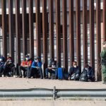 ‘Send Them Back’ Chants Erupt After ‘Strictest-Ever’ Migration Law PASSED! Group of people sitting by a tall border fence with an officer standing nearby
