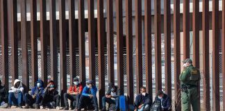 Group of people sitting by a tall border fence with an officer standing nearby