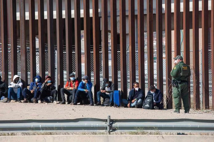 shutterstock_2079408472 Group of people sitting by a tall border fence with an officer standing nearby