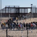 Crowd of people lining up near a border fence with barbed wire