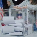 Trump FIRES Key Officials Weeks Before Midterms! Person casting a ballot into a clear box at a polling station