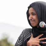 Ilhan Omar speaking at a microphone with hand on chest