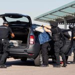 ICE Ambush Ends With Crushed Fed Agent customs officers detain a person beside a car with its trunk open