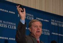3 Male Hookers Claim Lindsey Graham Paid For Sex Man in suit speaking at podium holding up a smartphone